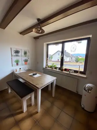 Heller Essplatz mit Holztisch und Bank vor großem Fenster, Kräuterbilder an der Wand und Blick ins Grüne.