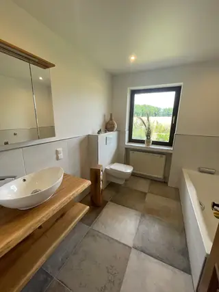Modernes Tageslichtbad mit Holzwaschtisch, Badewanne, wandhängendem WC und Fenster mit Feldblick.
